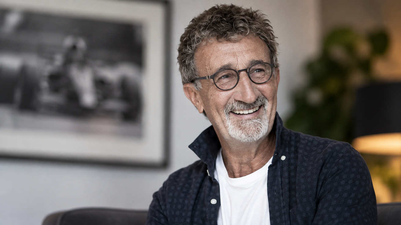 Homme souriant avec lunettes et barbe poivre et sel, assis dans un intérieur avec une photo floue en arrière-plan.